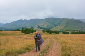 Fototapeta premium Satisfied tourist returns from mountains in overcast. Happy man in raincoat walks through hills and forest in bad weather. Traveler goes towards adventure. Hiker and mountain range under cloudy sky.