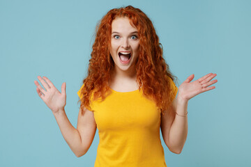 Young shocked surprised amazed redhead woman 20s wearing yellow t-shirt look camera spread hands say wow great isolated on plain light pastel blue background studio portrait. People lifestyle concept.