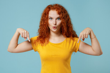 Young smiling happy redhead woman 20s in yellow t-shirt look camera point index finger down on workspace area mock up isolated on plain light pastel blue background studio People lifestyle concept