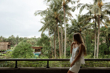 Naklejka premium Young beautiful slender girl stands on the balcony against the backdrop of palm trees