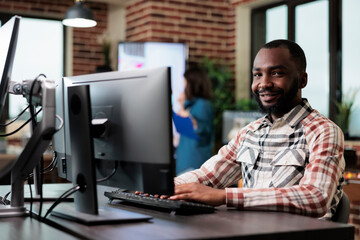 Agency adult happy smiling worker at computer in startup studio space modern interior. African american creative company employee sitting at desk in office workspace while looking at camera.