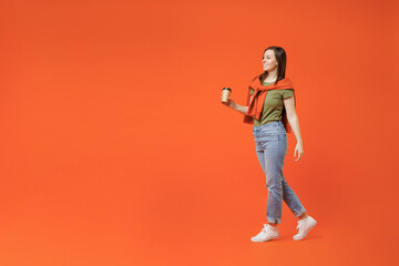 Fototapeta na wymiar Full body side view young happy fun woman 20s in khaki t-shirt tied sweater on shoulders hold takeaway delivery craft paper brown cup coffee to go walk go isolated on plain orange background studio