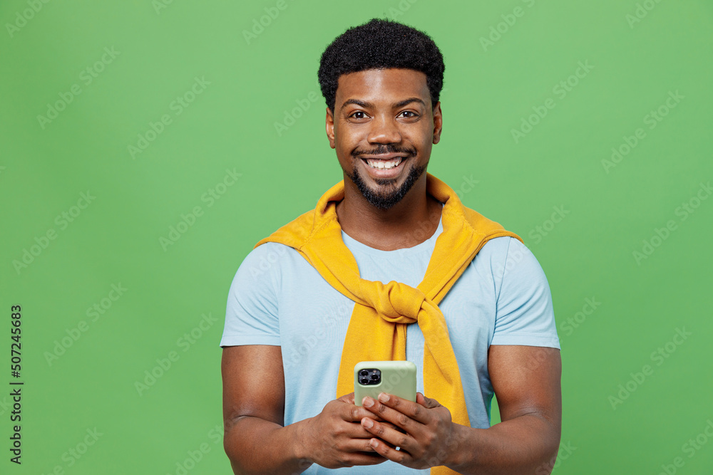 Wall mural Young man of African American ethnicity 20s wear blue t-shirt hold in hand use mobile cell phone chatting browsing internet isolated on plain green background studio portrait People lifestyle concept. - Wall murals