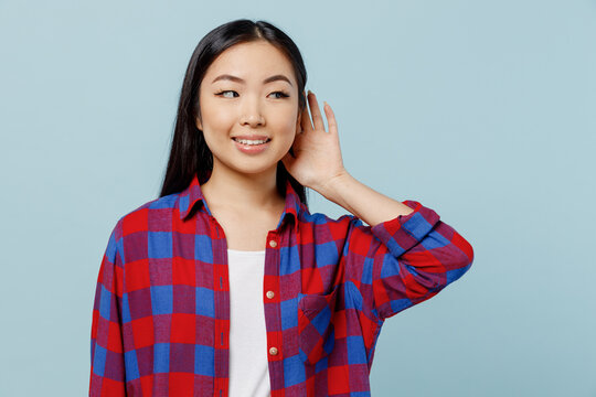 Young smiling curious nosy woman of Asian ethnicity 20s wear checkered shirt try to hear you overhear listening intently isolated on plain pastel light blue color background. People lifestyle concept.