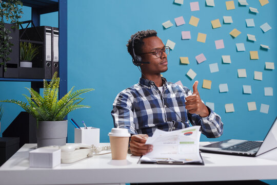 Helpline center operator wearing headset, showing thumbs up approval sign while on call with customer. Support service representative in conversation with client while looking over management charts.