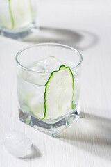 Selective focus of glass of cold refreshing vegetarian cucumber water or juice garnished or decorated with vegetable slice served with ice cubes on white wooden background with shadow as summer drink 