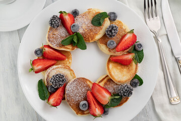 Appetizing pancakes with slices of strawberries, blueberries, blackberries on a white plate, cutlery on a light table.Homemade food.