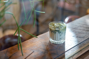A glass of Iced matcha latte green tea with ice and milk on a wooden bar over a cafe glass window reflex at a Cafe coffee shop. Cold brew refreshment summer drink with copy space. Selective focus