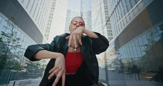 Female fashion model wearing black leather jacket short skirt and cool red eyeglasses dancing seductively near high rise glass buildings skyscrapers downtown. Urban style woman in modern city