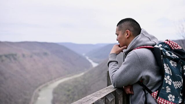 A boy whose hair is cut short wearing a pull over and carrying a bag on his back, standing on a bridge with his hand on his cheek looking at the mountain of New River Gorge, West Virginia