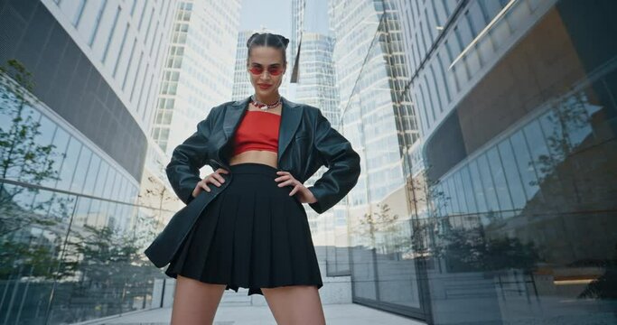 Stylish young woman wearing black jacket short skirt and cool red eyeglasses posing with hands on hips standing on stairs near high rise glass skyscrapers modern buildings. Urban style fashion model