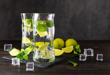 Two tall transparent glasses of mojito on a shale dark plank. Lime wedges and mint leaves in the background. Pieces of ice on a dark wooden table. Dark gray background, space for text