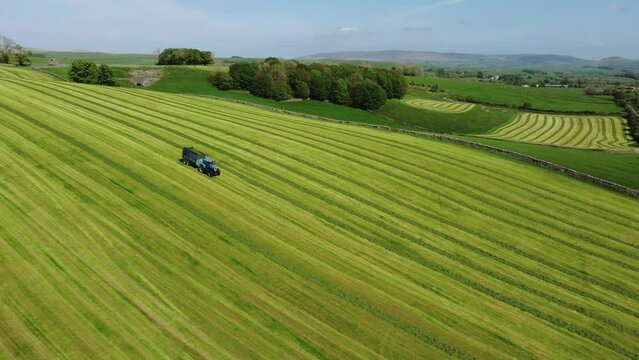 Tractor with trailer full of grass driving on green field hills in Yorkshire UK countryside. Drone aerial
