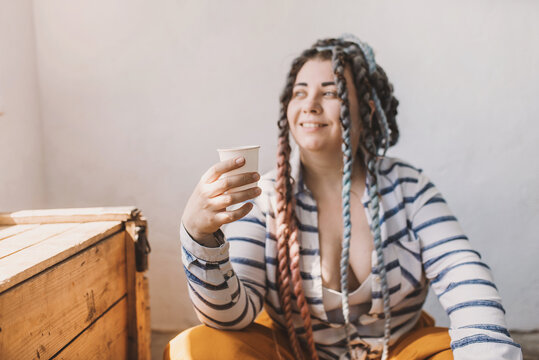 Very emotional happy positive plus size woman with dreadlocks in bright yellow pants in a modern bohemian boho interior holds a disposable paper cup in her hands and drinks coffee. Ecological approach