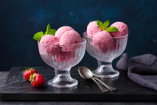 Strawberry ice cream in a glass bowl on marble cutting board. Dark background.