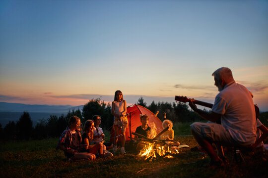 Happy children sitting around campfire, singing songs with guitar at dusk. Side view adult guide playing guitar for kids near fire in mountains, with colorful sky on background. Concept of camping.