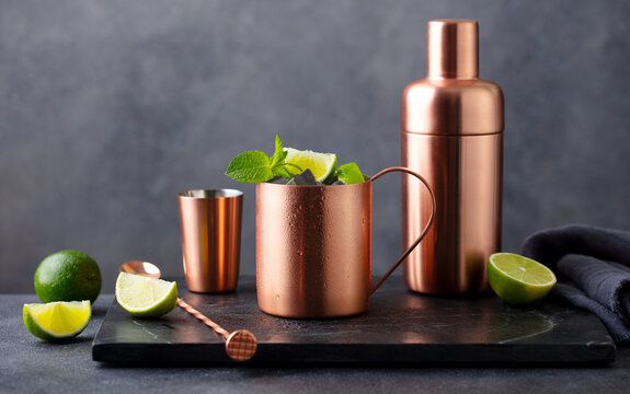 Moscow mule cocktail in copper mug, shaker with fresh mint, lime on marble board. Dark background. Copy space.