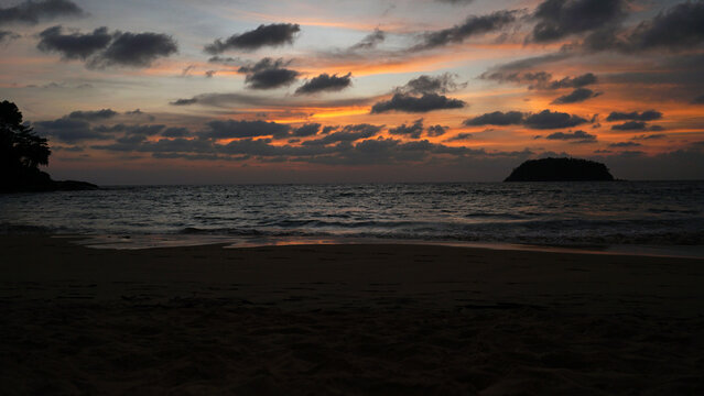 An epic sunset with a view of the sea and the island. The orange sky is covered with dark clouds. There is a lonely island in sea. Silhouettes of trees are visible. The sky is reflected on wet sand