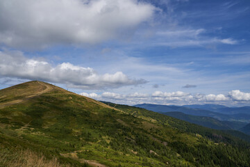 Scenic view of gentle slope mountain, with path leading to hilltop in summer. Cumulus clouds, crossing blue sky, seemed touching top of mount, with hilly landscape on background. Concept of highland.