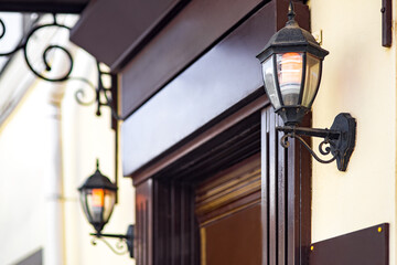 2 iron retro lanterns on the wall of the facade of the building at the entrance central door with wooden cladding of the front door, retro-style electric lamps with a warm glow of light, side view.