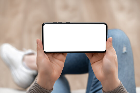 Top view mockup image of woman holding mobile phone horizontally with blank white screen. woman using cellphone horizontal with while sitting cross-legged on sofa at home. Mockup smartphone