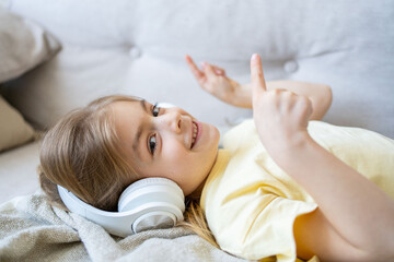 Joyful child school-aged girl listening to music at home, lying on sofa using wireless headset. home, technology and music concept.