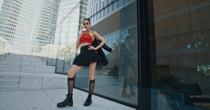 Sexy young woman wearing black jacket short skirt stockings and cool red eyeglasses posing standing on stairs by wall of high rise glass buildings downtown. Gorgeous cheeky urban style fashion female