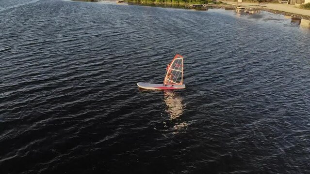 A novice windsurfer girl is learning to steer a windsurfing board with a sail on a lake at sunset