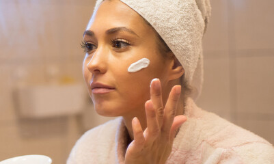 Young beautiful woman in bathrobe putting facial mask on face in front of the bathroom mirror