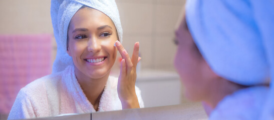 Fototapeta premium Young beautiful woman in bathrobe putting facial mask on face in front of the bathroom mirror