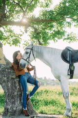 Woman stands near tree and holds white horse by the bridle.