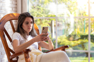 Woman using the smartphone sitting in a rocking chair on the window with garden in the background