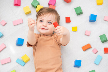 cute smiling baby girl in brown bodysuit laying on floor around colorful wooden blocks. top view