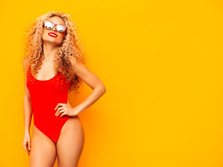 Young beautiful smiling woman posing near yellow wall in studio.Sexy model in red swimwear bathing suit.Positive female with curls hairstyle.Happy and cheerful. In sunglasses