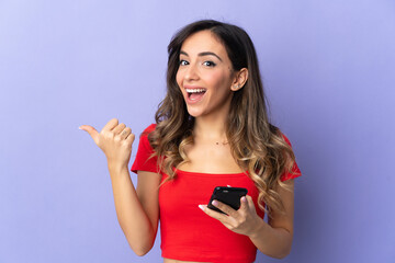 Young caucasian woman isolated on purple background using mobile phone and pointing to the lateral