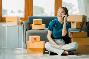 A young Asian woman starts a business to talk to customers via smartphone. Work happily. with box and laptop at home Prepare to deliver parcels in SMEs, supply chains, online procurement concepts.