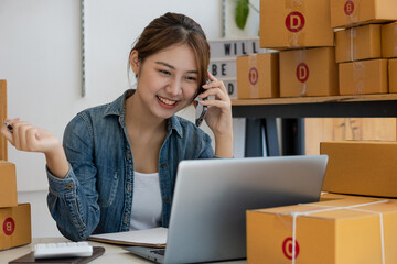 A young Asian woman starts a business to talk to customers via smartphone. Work happily. with box and laptop at home Prepare to deliver parcels in SMEs, supply chains, online procurement concepts.