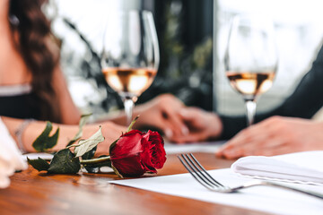 Romantic Relationship And Celebration. Selective focus on red rose. Couple having dinner, holding hands in blurred background. Young couple in love spending time together at home or cafe - obrazy, fototapety, plakaty