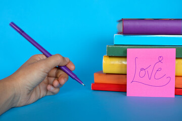 horizontal photograph with a hand holding a pen after writing love on a postit leaning on the spines of books piled up forming the lgtb flag. respect for sexual orientation.