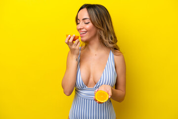 Young caucasian woman isolated on yellow background in swimsuit and holding an orange