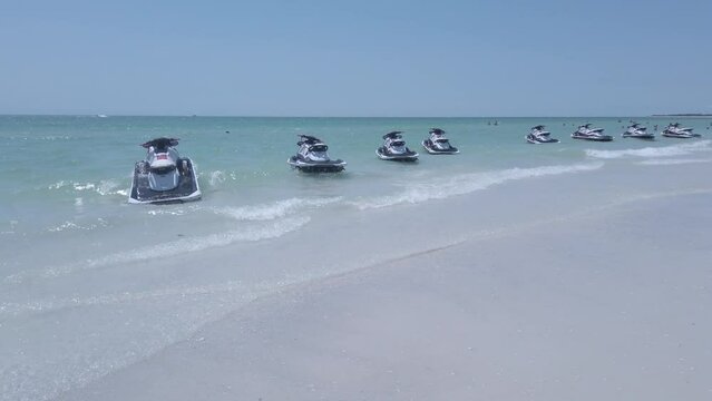 A row of wave runners are seen bobbing in beautiful clear ocean water near a soft white beach 
