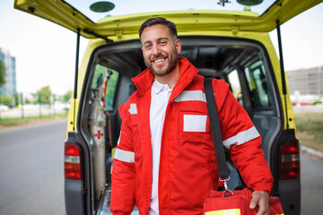 Fototapeta na wymiar Young man , a paramedic, standing at the rear of an ambulance, by the open doors. He is looking at the camera with a confident expression, smiling, carrying a medical trauma bag on his shoulder.