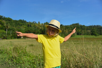 The preschooler shows the direction with his hand. A little curly-haired boy walks in nature in a hat. Vacation, traveler, trip, summer time. Green grass and blue sky in the background, lifestyle