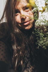 Beautiful sensual woman portrait in sunlight among summer wildflowers in room. Young brunette woman with flowers posing in sunlight. Skin care and self care