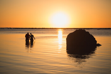 Two men fishing in the ocean from the beach at sunset
