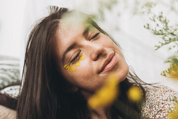 Beautiful sensual woman with yellow petals on face posing with wildflowers in sunny room. Portrait of young brunette in boho dress  with flowers. Skin care and self care. Summer mood