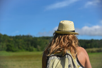 A curly-haired man walks in nature in a hat and with backpack. Vacation, traveler, trip, summer time. Green grass and blue sky in the background, lifestyle, back view