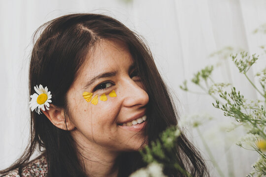 Beautiful happy woman with yellow petals under eyes posing with wildflowers in sunny room. Portrait of young brunette female in boho dress smiling with flowers. Skin care and self care. Summer mood