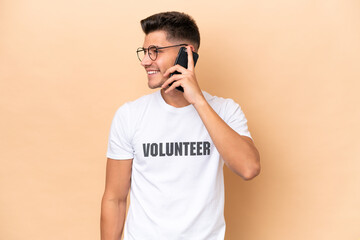 Young volunteer caucasian man isolated on beige background keeping a conversation with the mobile phone with someone