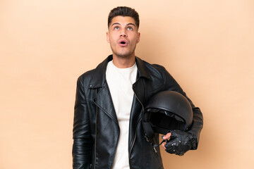 Young caucasian man with a motorcycle helmet isolated on beige background looking up and with surprised expression